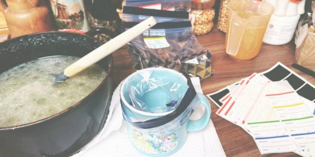 A pot of home-made broth with a wooden spoon on a kitchen counter, beside storage jars, a covered cup and freezer labels